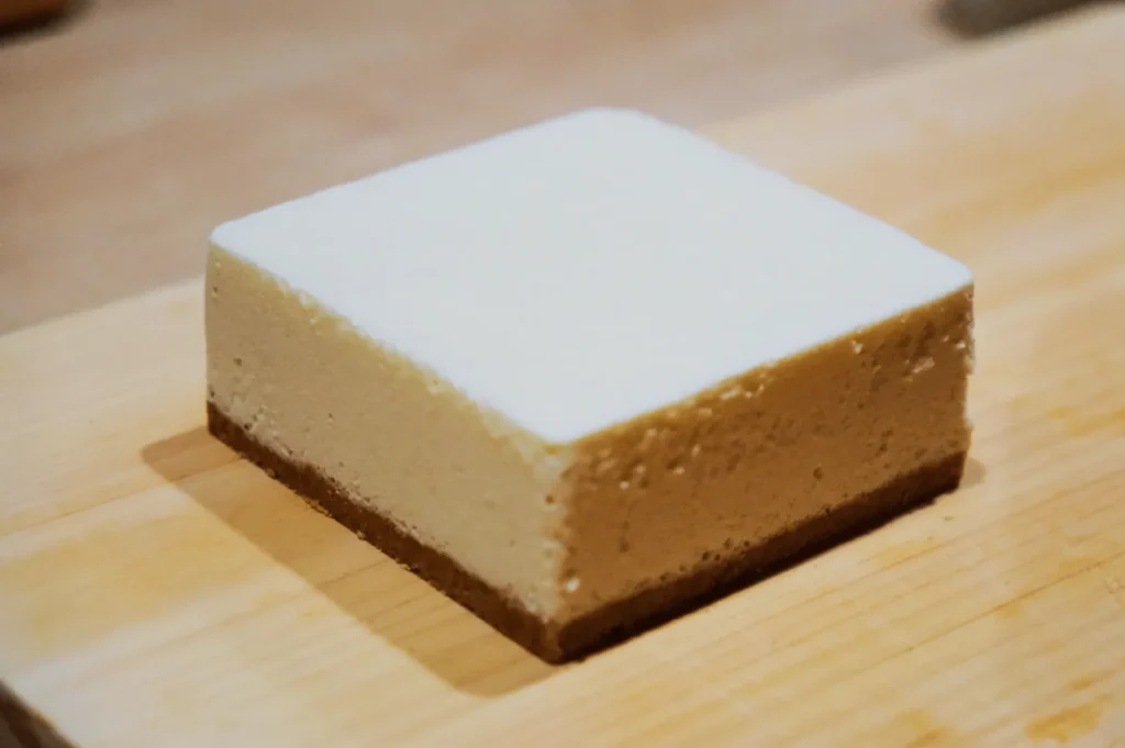 A rectangular slice of creamy white cheesecake with a light brown graham cracker crust, placed on a wooden surface, conveying a smooth and indulgent treat.