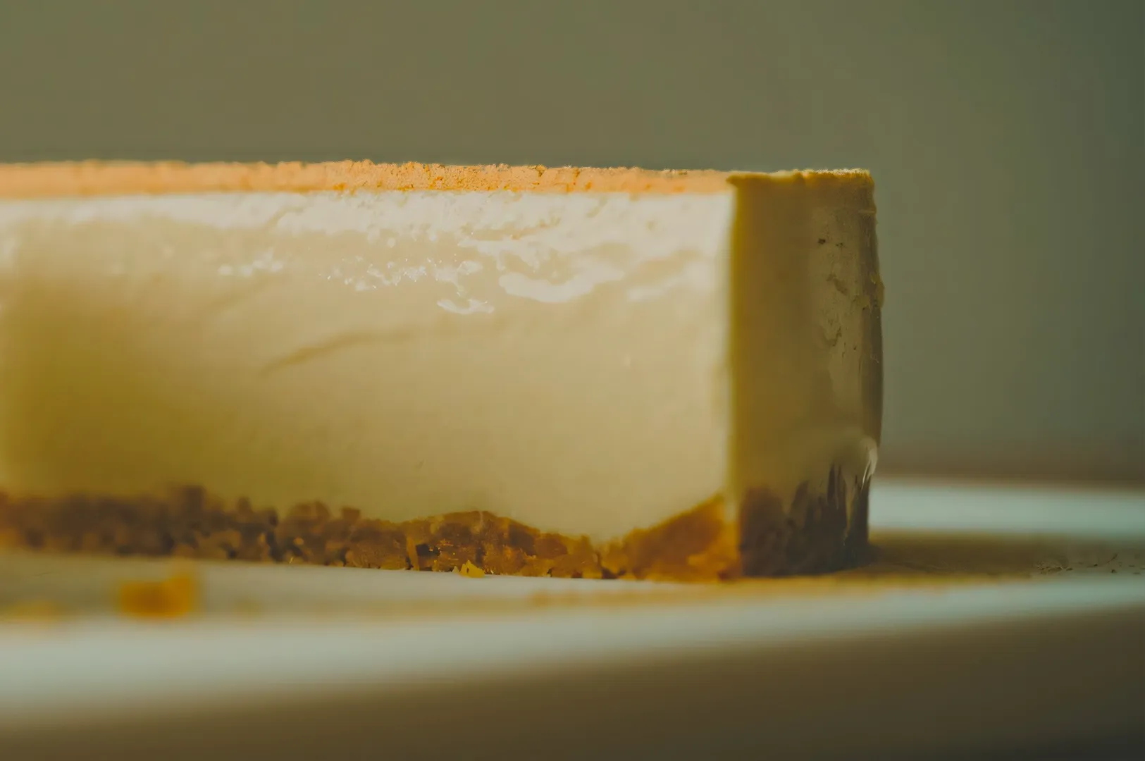 Close-up of a cheesecake slice with a smooth creamy top and a golden-brown crumbly crust on a white plate. The lighting highlights its glossy texture.