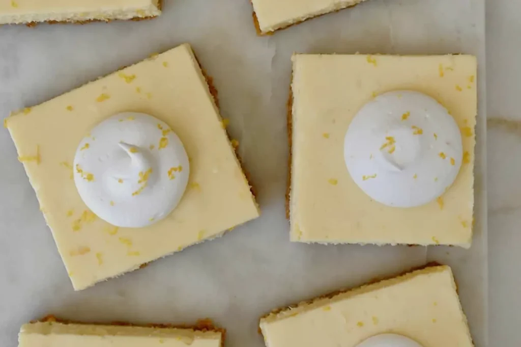 Top view of lemon bars with a creamy topping. The bars are square with a light yellow hue, sprinkled with zest, evoking a fresh, tangy feel.