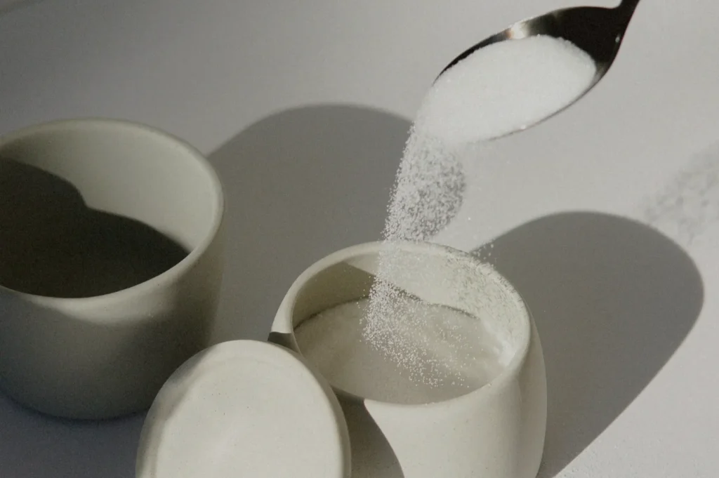 A spoon pours white sugar into a small ceramic container with a lid beside it. The scene is softly lit, casting gentle shadows on a light background.