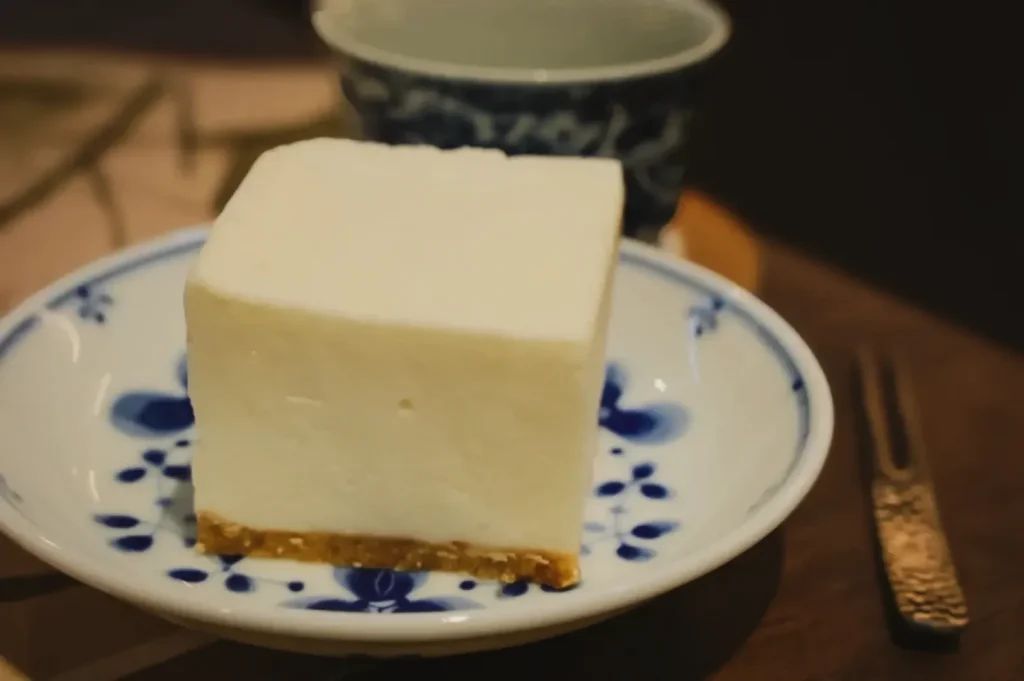 A creamy, white tofu cheesecake slice with a light brown crust sits on a blue and white floral plate. A blue-patterned cup and a fork are nearby.