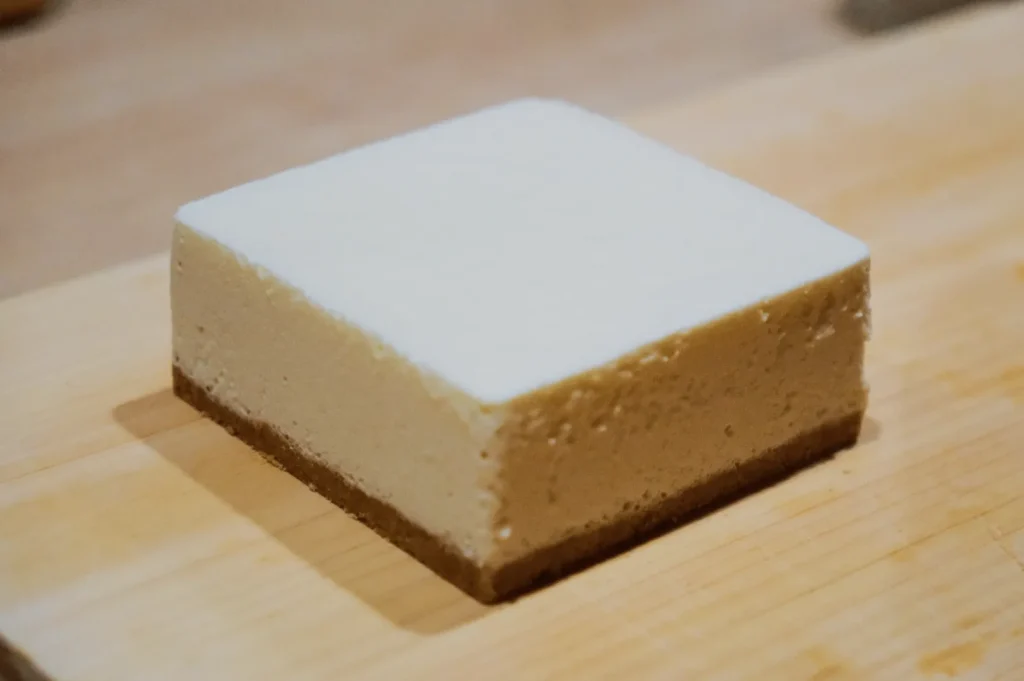 A neat square slice of cheesecake with a smooth cream layer on a light brown crumb crust, placed on a wooden board, conveying a simple and inviting look.