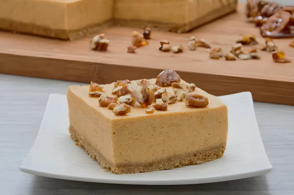 A square slice of pumpkin cheesecake, topped with chopped nuts, sits on a white plate. More cheesecake and nuts are visible in the background on a wooden board.