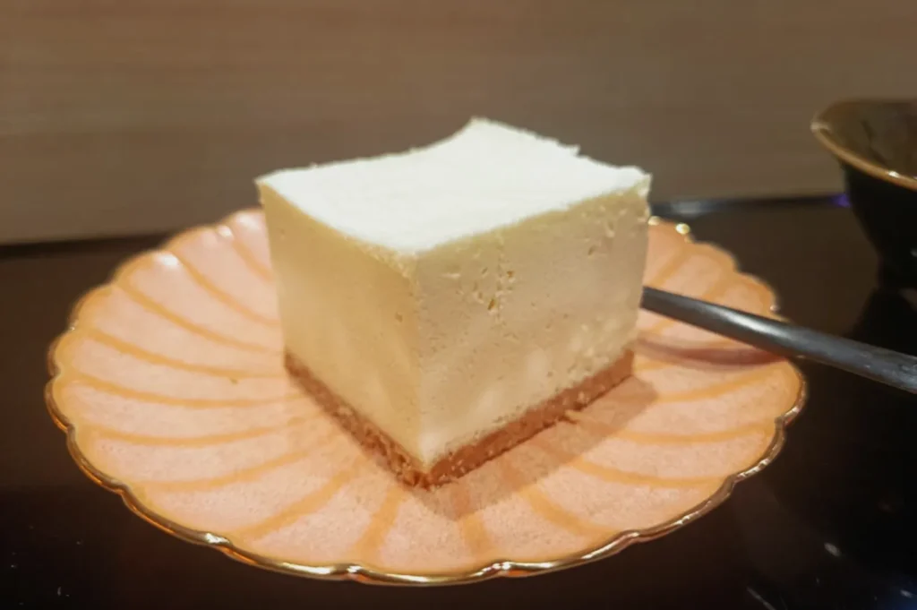 A slice of creamy cheesecake with a golden brown crust sits on a pink scalloped plate, next to a silver spoon, on a reflective surface.
