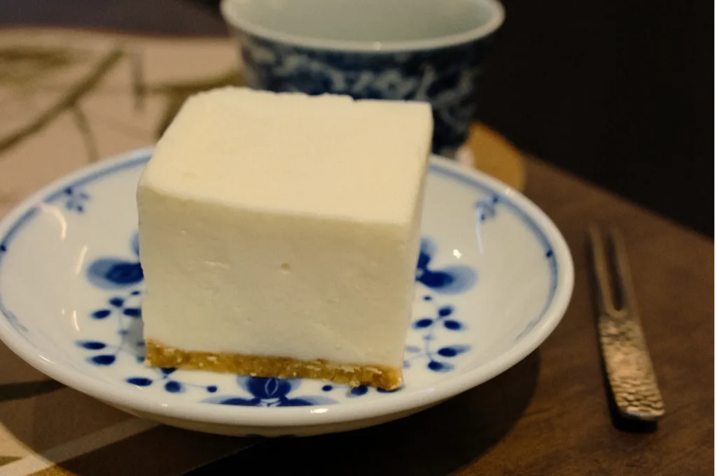 A square slice of creamy cheesecake on a blue and white floral plate beside a textured fork. A patterned cup is in the background, creating a cozy atmosphere.
