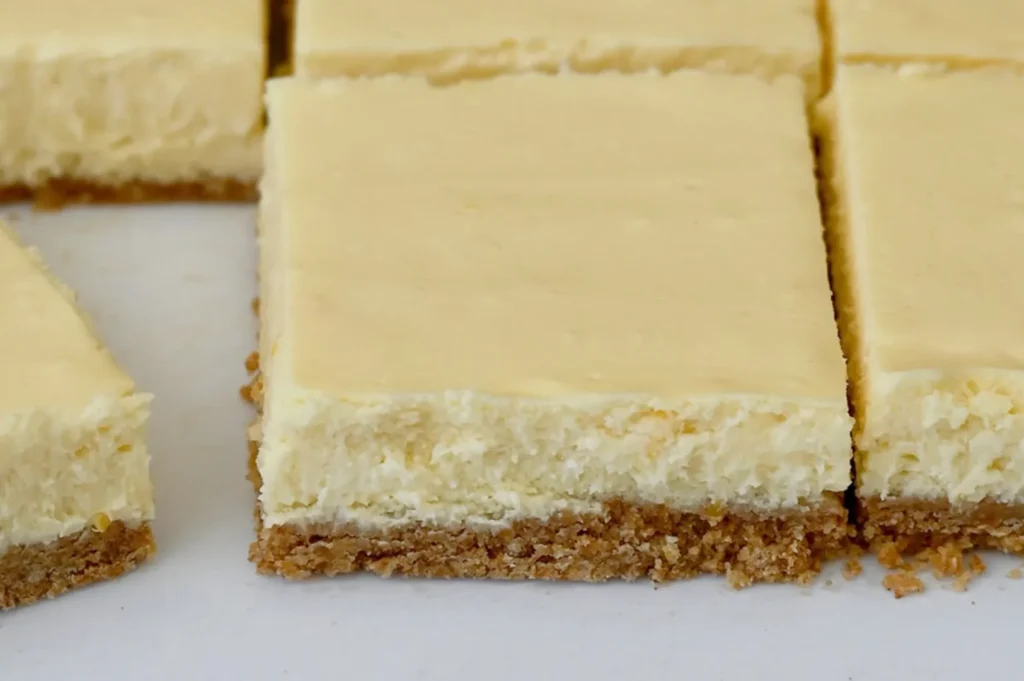 Close-up of creamy, square-shaped cheesecake bars with a golden graham cracker crust, cut into neat sections on a white surface. The texture looks rich.