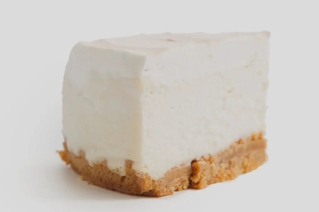 A slice of creamy cheesecake with a golden-brown biscuit crust on a white background. The texture looks smooth and rich, suggesting indulgence.