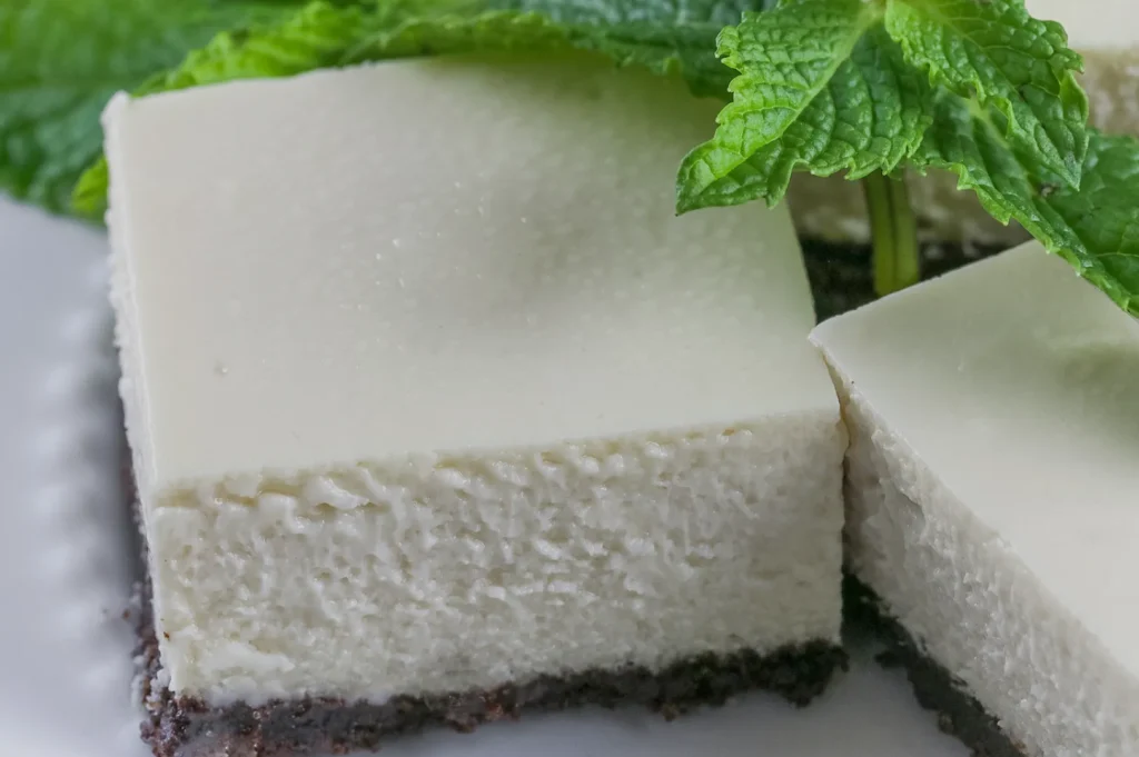 Close-up of creamy lime cheesecake bars with a chocolate crust, garnished with fresh mint leaves. The texture appears smooth and inviting.