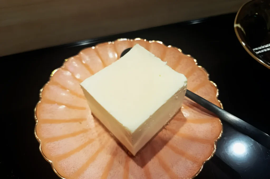 A square piece of creamy white dessert sits on a scalloped pink plate with a spoon beside it. The setting is elegant and inviting.
