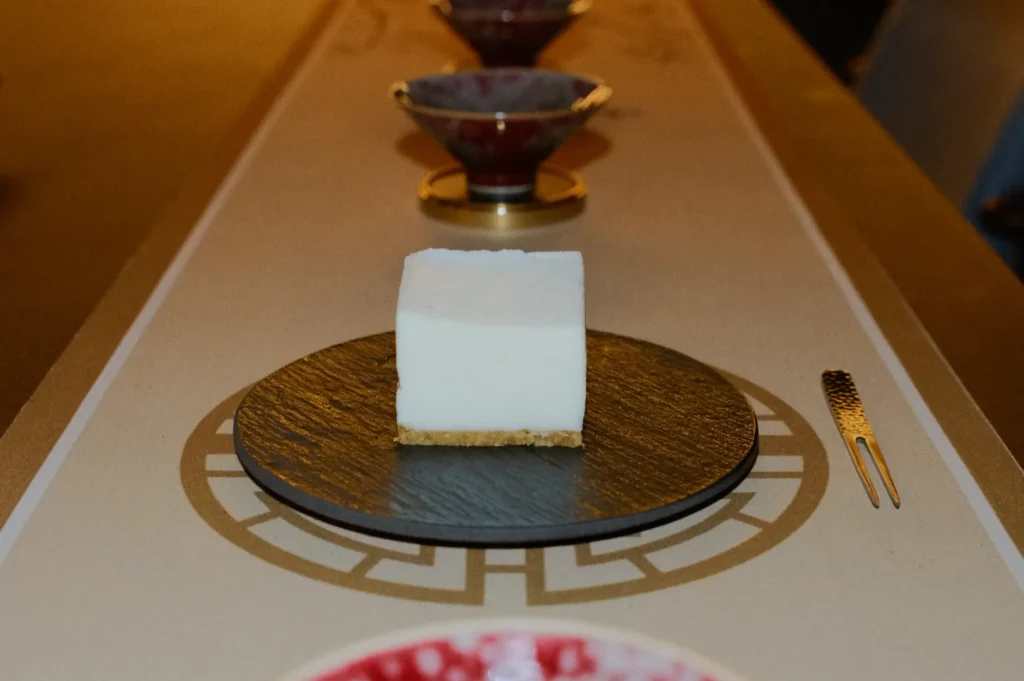 A perfectly square, white dessert is centered on a slate plate on an elegant table. Behind it, patterned bowls suggest a refined dining setting.