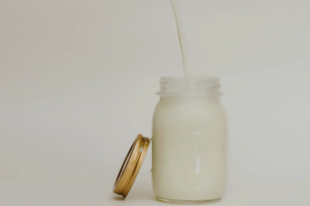A stream of milk pours into a clear mason jar beside its golden lid on a plain white background. The scene conveys simplicity and freshness.