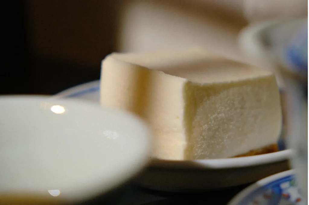 A soft, square piece of silky tofu sits on a small plate. The tofu's creamy texture contrasts with the dark, blurred background, creating a serene and simple aesthetic.