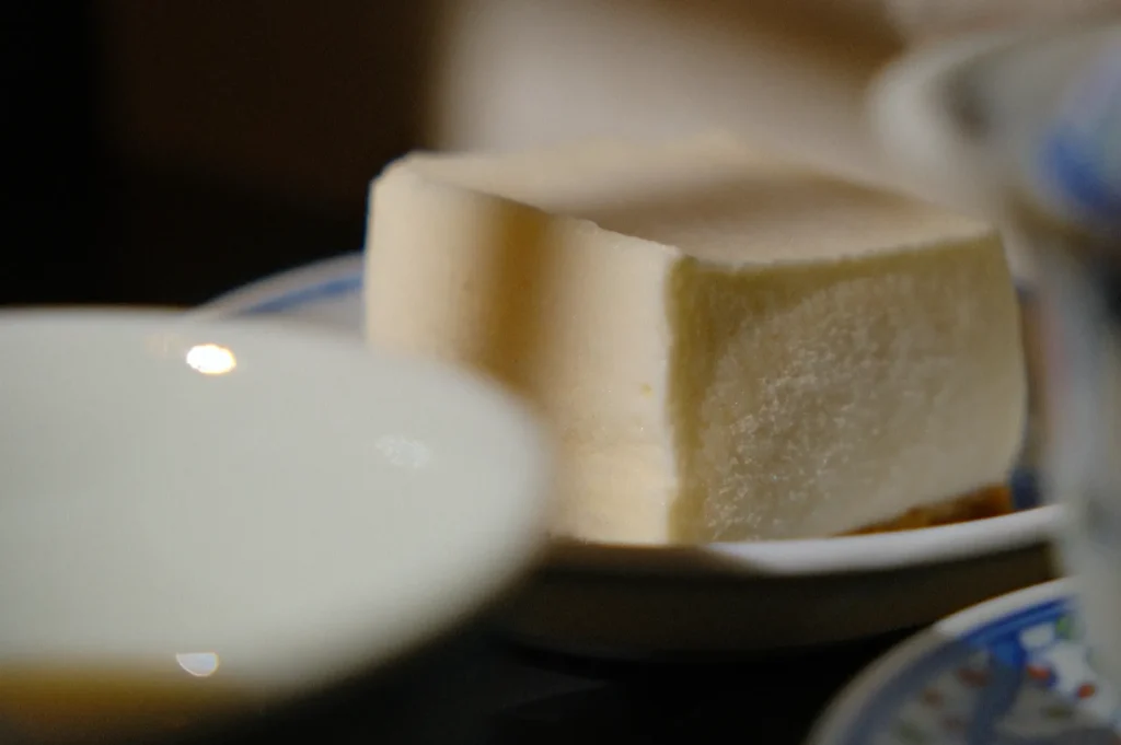 A close-up of a creamy, rectangular piece of tofu on a patterned plate, surrounded by blurred dishes, creating a warm and cozy dining atmosphere.