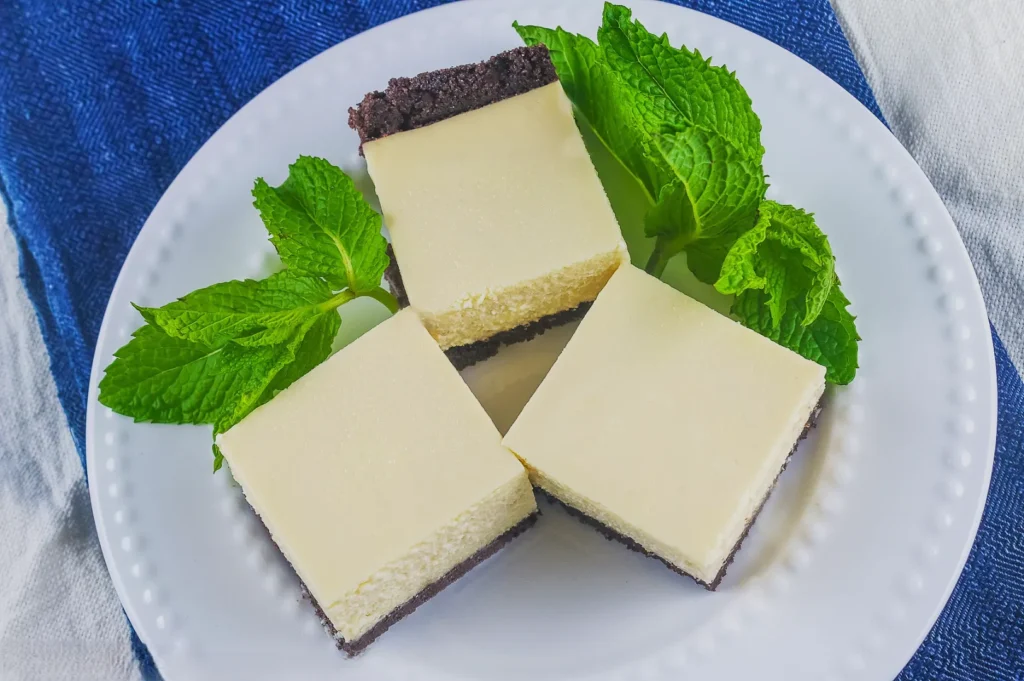 Three creamy cheesecake squares with a chocolate crust are on a white plate, garnished with fresh mint leaves, set on a blue cloth.