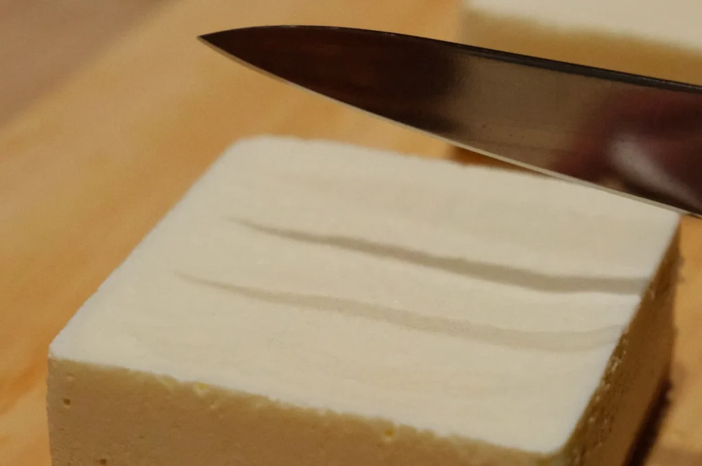 A knife poised above a block of tofu on a wooden cutting board, conveying a sense of meal preparation with focus on the tofu's smooth texture.