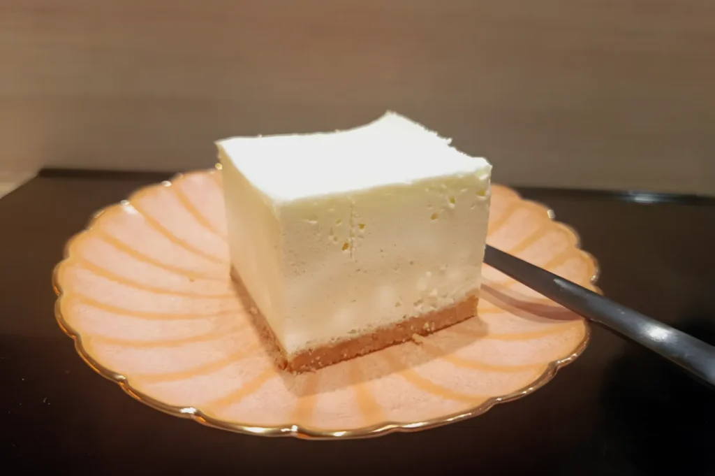 A square slice of white cheesecake with a thin crust sits centered on a delicate pink scalloped plate. A spoon rests beside it, conveying a sense of indulgence.