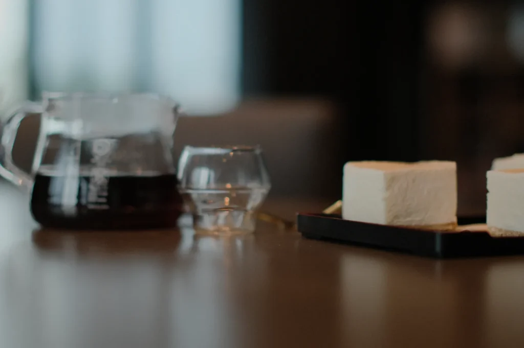 A glass pitcher of black coffee sits beside an empty glass on a wooden table. Nearby, square pieces of cake rest on a black tray. The scene feels cozy and inviting.