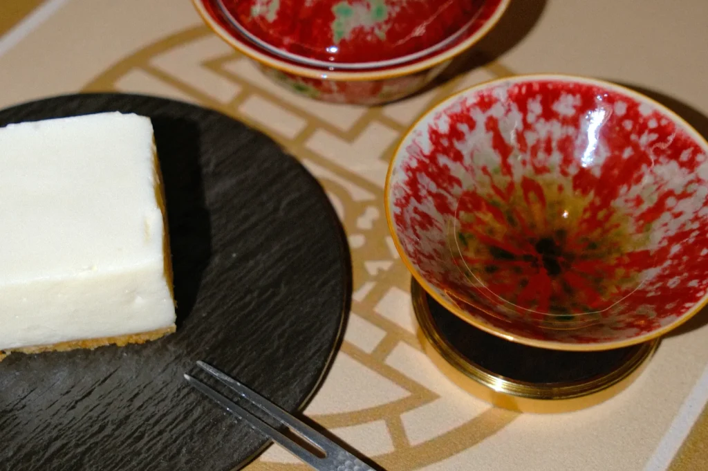 A small white cheesecake slice on a round black plate with a fork beside it. Two red and gold speckled bowls are nearby on a patterned surface.
