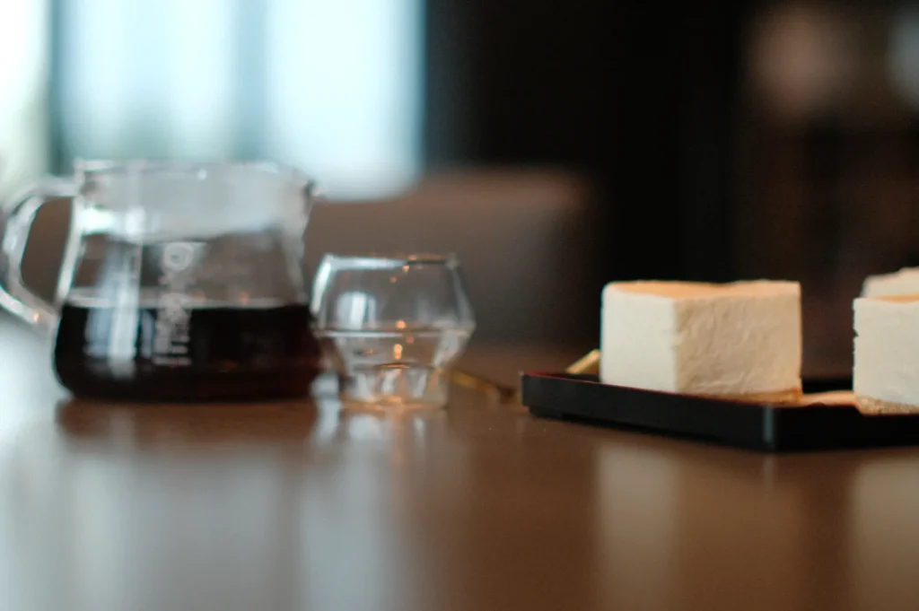 A glass carafe of dark coffee and a small cup sit on a wooden table, next to a black tray holding two cubes of soft, white cake. The setting feels calm and cozy.