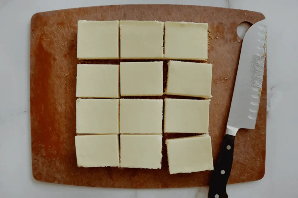 Twelve creamy cheesecake squares neatly arranged on a brown cutting board, with a knife covered in crumbs placed on the right side, creating a tidy and inviting look.