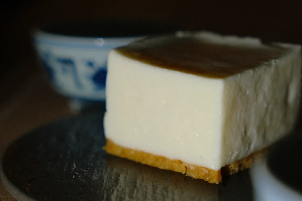 A slice of creamy cheesecake with a golden brown top and a crust sits on a dark plate. A blurred, patterned bowl is in the background.