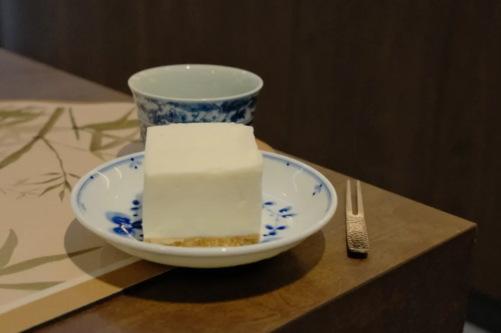 A creamy white dessert square on a blue floral dish beside a decorative teacup and fork, placed on a wooden table with a bamboo-patterned placemat.
