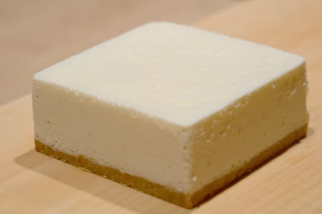 A neatly cut square slice of cheesecake sits on a wooden surface. The dessert has a smooth cream-colored top and a thin, darker crust base.