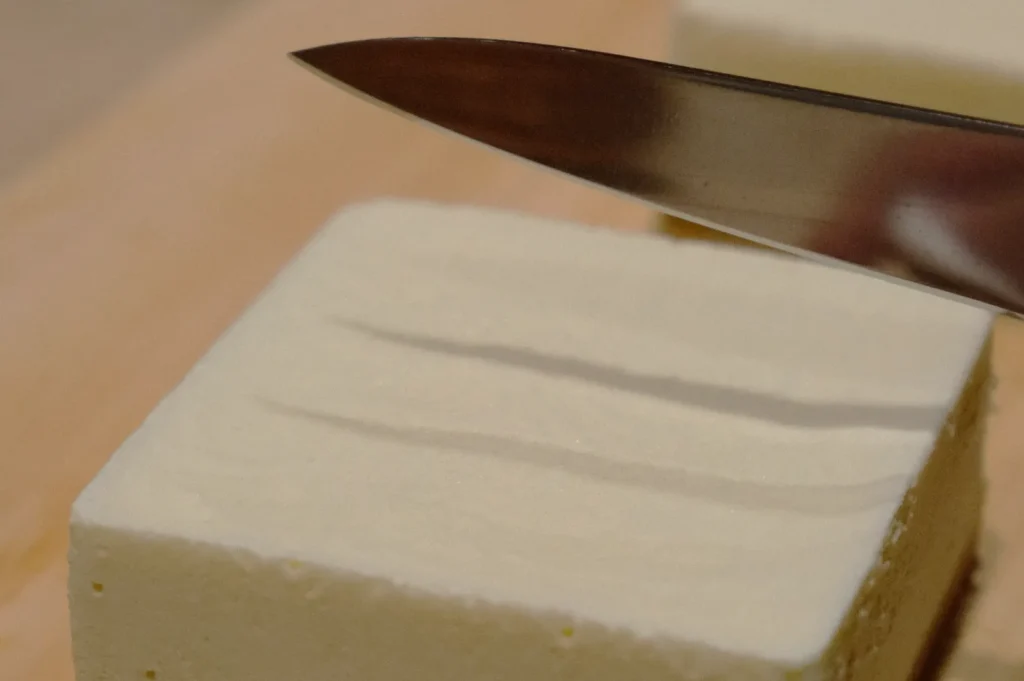 Close-up of a knife slicing into a smooth, white block of tofu on a wooden cutting board. The scene conveys a sense of precision and preparation.