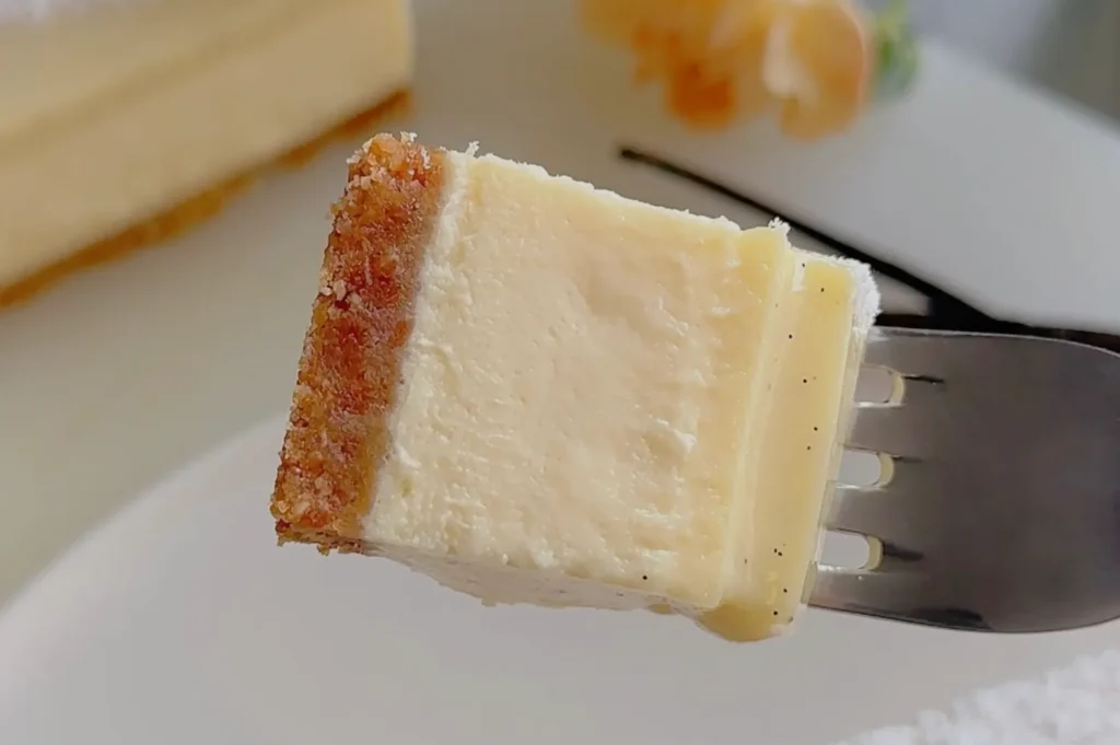 A fork holds a creamy slice of cheesecake with a golden-brown crust. The background shows a larger cheesecake and a soft-focus decorative flower.