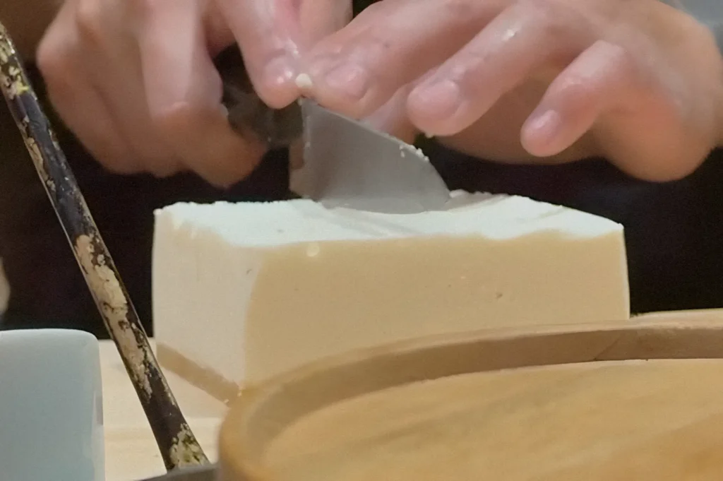 Hands carefully slice a block of tofu with a knife on a wooden board. The setting conveys a calm, focused atmosphere, highlighting culinary preparation.