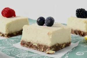 Three creamy cheesecake squares on a green patterned cloth, each topped with berries: raspberry, blueberries, and blackberry, on a nutty crust.