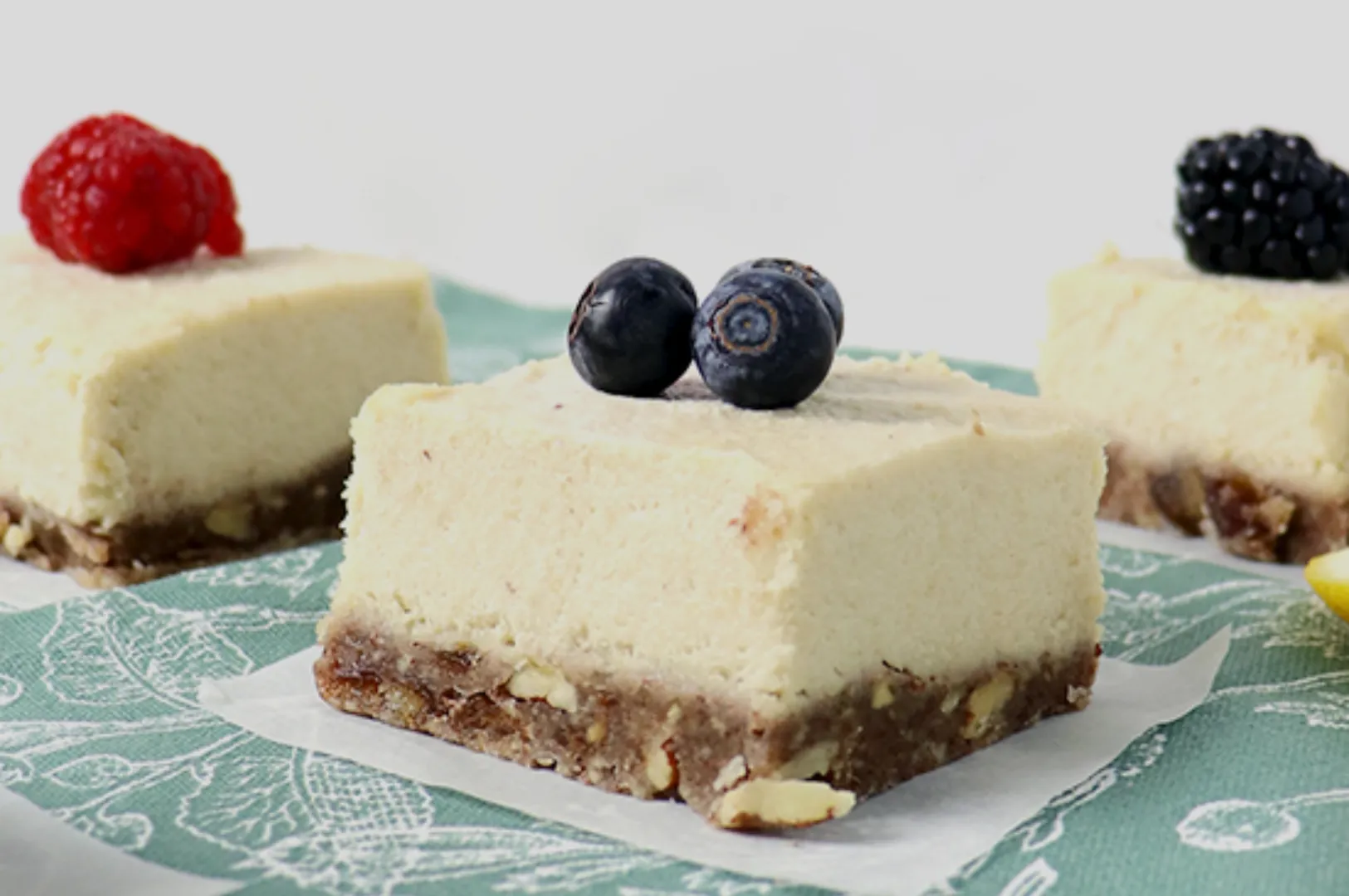 Three creamy cheesecake squares on a green patterned cloth, each topped with berries: raspberry, blueberries, and blackberry, on a nutty crust.