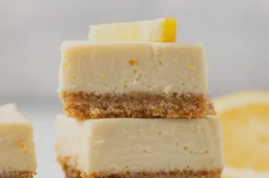 Two square lemon bars stacked, featuring creamy yellow tops and a crumbly brown crust. A lemon slice garnishes the top, in a bright, fresh setting.