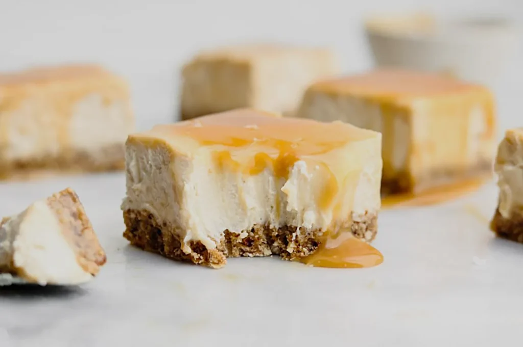 Decadent cheesecake bars topped with caramel sauce on a white surface. Smooth, creamy texture with a crumbly brown crust, creating an indulgent feel.