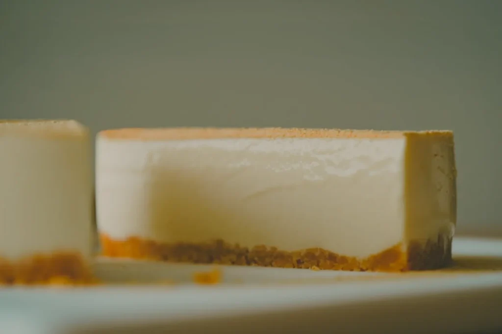 A creamy cheesecake slice with a graham cracker crust sits on a white plate. Soft lighting emphasizes its smooth texture and inviting appearance.