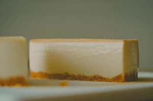 A creamy cheesecake slice with a graham cracker crust sits on a white plate. Soft lighting emphasizes its smooth texture and inviting appearance.