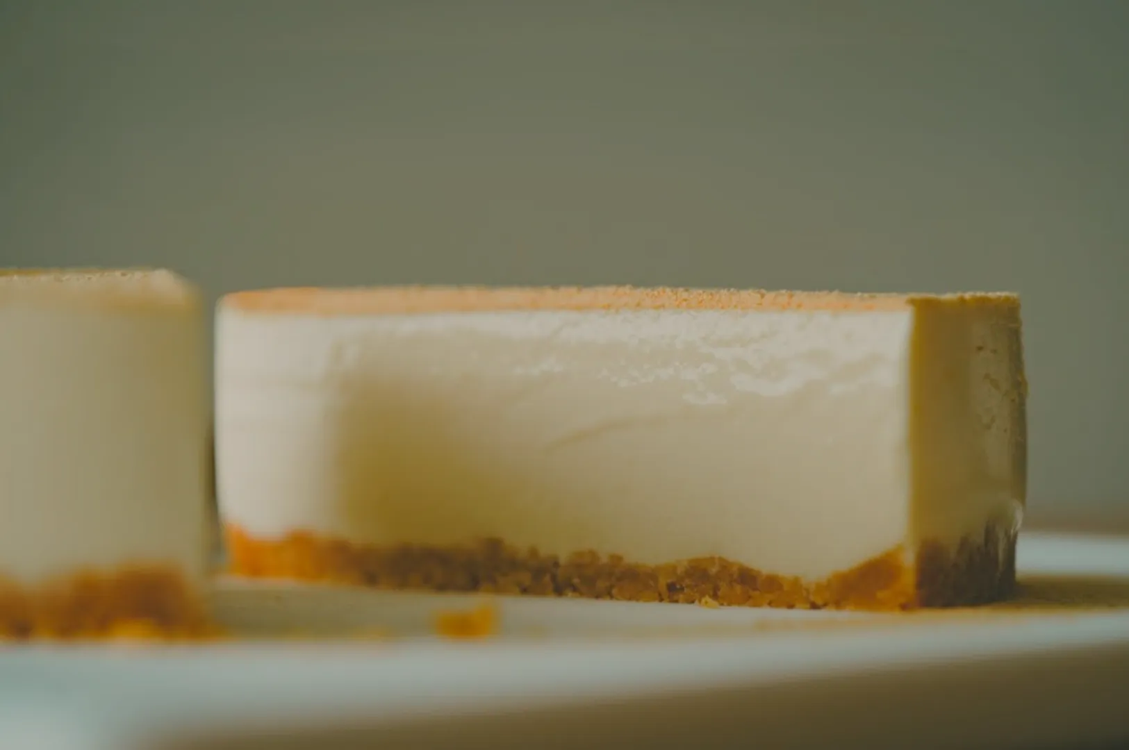 A creamy cheesecake slice with a graham cracker crust sits on a white plate. Soft lighting emphasizes its smooth texture and inviting appearance.
