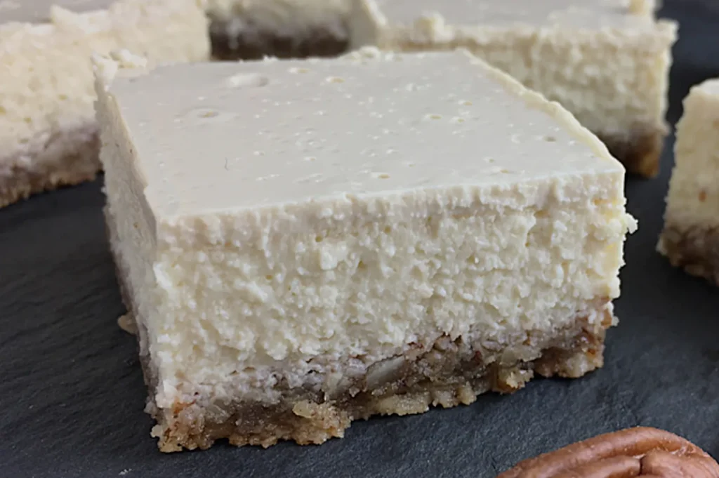 A close-up of a creamy cheesecake bar with a textured, pale top layer and a crumbly, nutty brown crust on a dark slate surface.