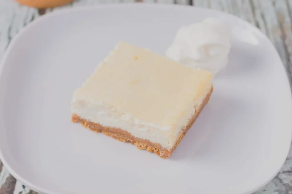 A square slice of creamy cheesecake on a white plate, featuring a smooth, pale yellow top and a brown crust base, garnished with whipped cream.