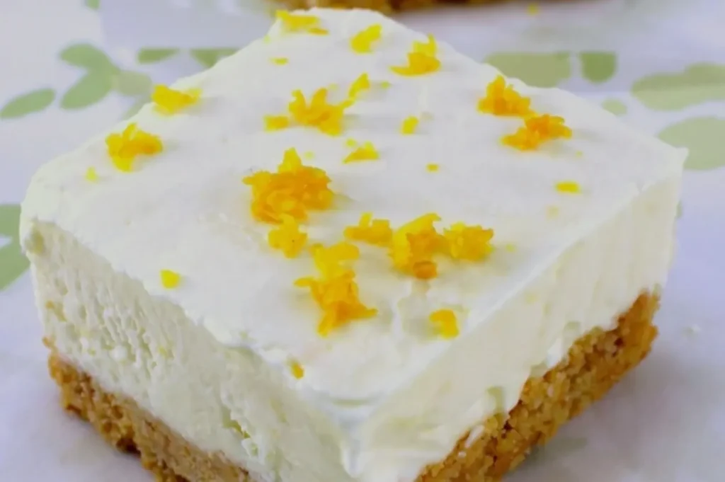 A creamy lemon dessert square with a graham cracker crust, topped with fluffy lemon filling and sprinkled with lemon zest, on a white plate.