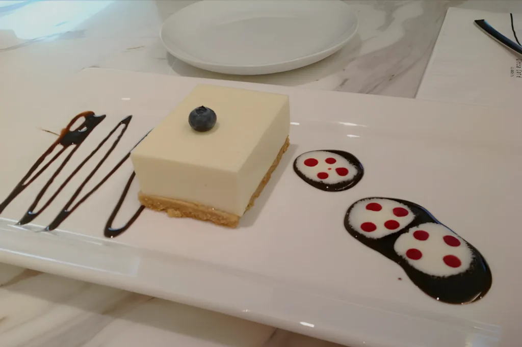 A square cheesecake with a blueberry on top is presented on a white plate. Decorative chocolate and cream swirls with red dots flank the dessert.