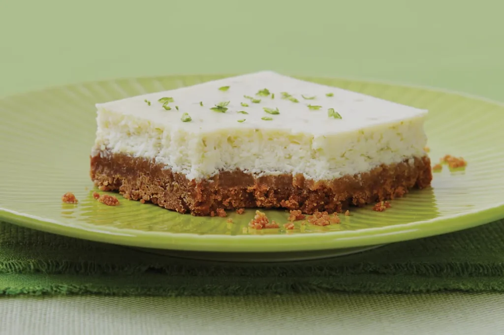 Close-up of a slice of lime cheesecake on a green plate. The dessert has a creamy top with lime zest, and a crumbly brown crust. Fresh and inviting.