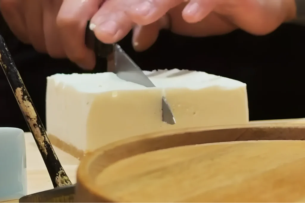Hands slicing a smooth, creamy cheesecake into neat portions on a wooden board. The setting feels cozy, focusing on the precise cut.
