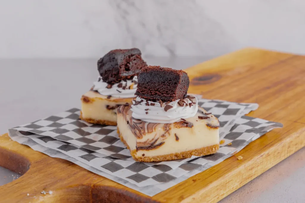 Two marbled cheesecake squares topped with whipped cream and chocolate brownie cubes sit on a checkered paper on a wooden board.