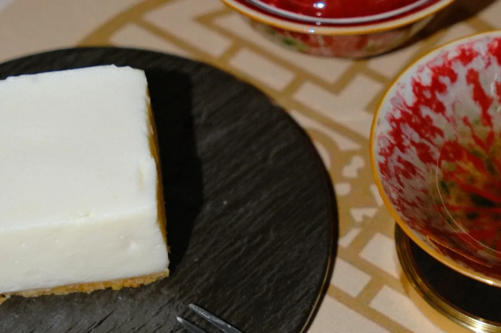 A slice of cheesecake on a dark plate, set on a patterned tablecloth. Nearby are colorful, decorative bowls, creating a cozy, elegant atmosphere.
