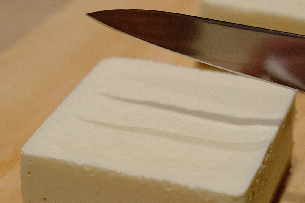 Close-up of a knife slicing firm tofu on a wooden board. The tofu is rectangular and smooth, suggesting a precise and careful preparation.