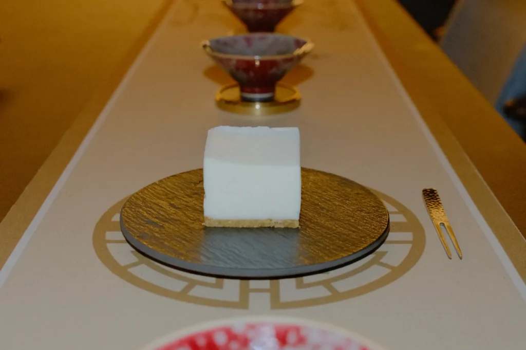 A square white dessert on a black plate, placed on a decorative table with a small fork beside it. Red bowls are arranged in a neat row behind.