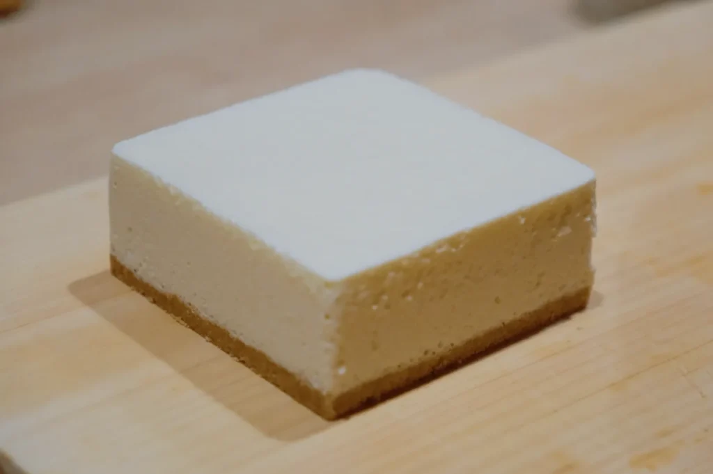 A neatly sliced square cheesecake rests on a wooden board. It has a creamy, pale yellow middle layer and a light brown crust, exuding a fresh and tempting appeal.