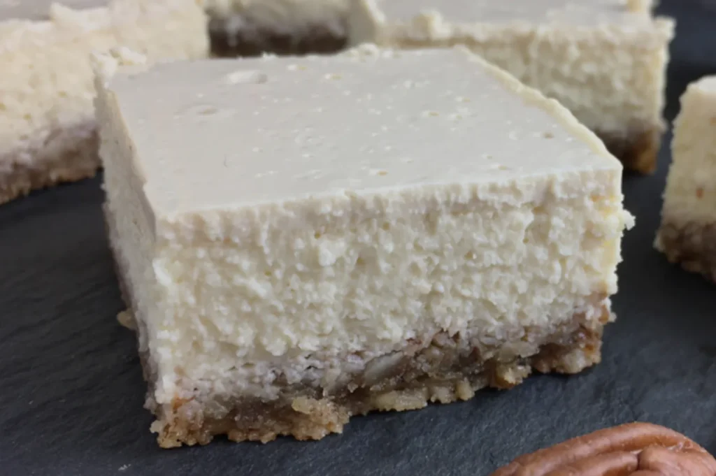 A close-up of a creamy, square cheesecake slice with a crumbly brown crust on a dark surface, evoking a rich and indulgent texture.