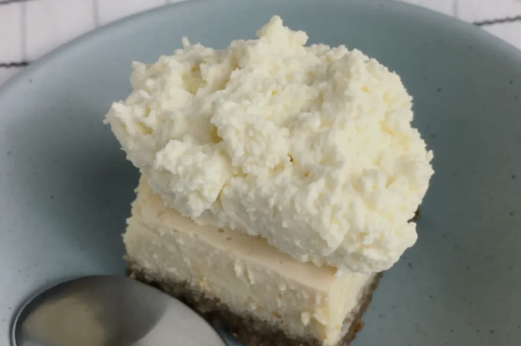 A slice of creamy cheesecake with a crumbly crust, topped with a thick layer of whipped cream, sits on a light blue plate beside a spoon.